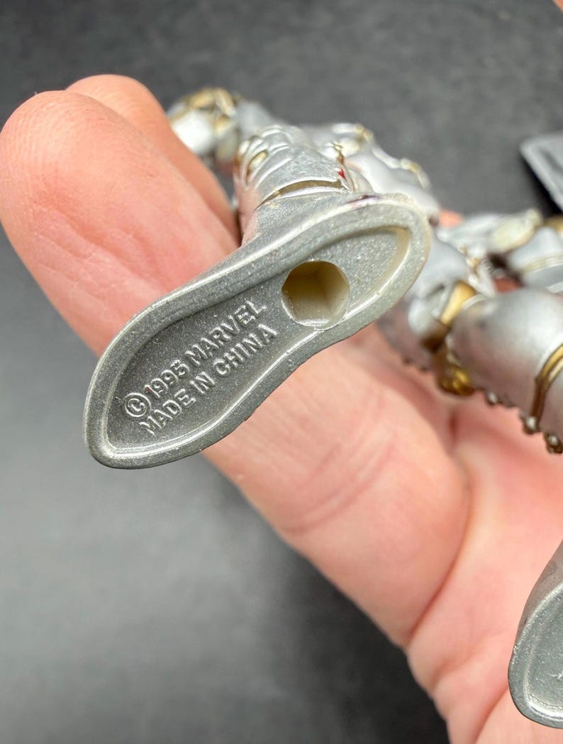 May include: A silver-colored action figure with gold accents. The figure's foot is visible, with the text "&copy;1995 MARVEL MADE IN CHINA" imprinted on the sole. The figure is held in a hand.