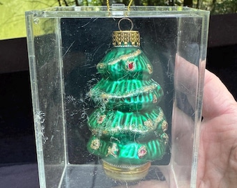 May include: A green Christmas tree ornament with gold and red accents, displayed in a clear acrylic box. The ornament has a gold cap and a gold string for hanging. The text "Hand Crafted Glass Ornament" is visible on the box.