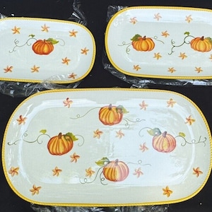 May include: Three white ceramic serving platters with a yellow rim and a pumpkin design. The platters are different sizes and feature orange pumpkins with green vines and leaves.