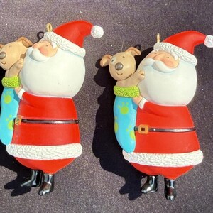 May include: Two Christmas tree decorations. Each features a Santa Claus figure in a red suit with white trim, holding a puppy in a blue stocking. The puppy is tan, and the stocking has green and blue accents.