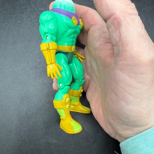 Op de afbeelding: Een groen actiefiguur met een bolvormig hoofd, paarse hoofdband en gele accenten op handschoenen, laarzen en riem. Het figuur wordt in een hand gehouden, wat de grootte en details laat zien. Het speelgoed heeft een gespierde lichaamsbouw en een vintage esthetiek.