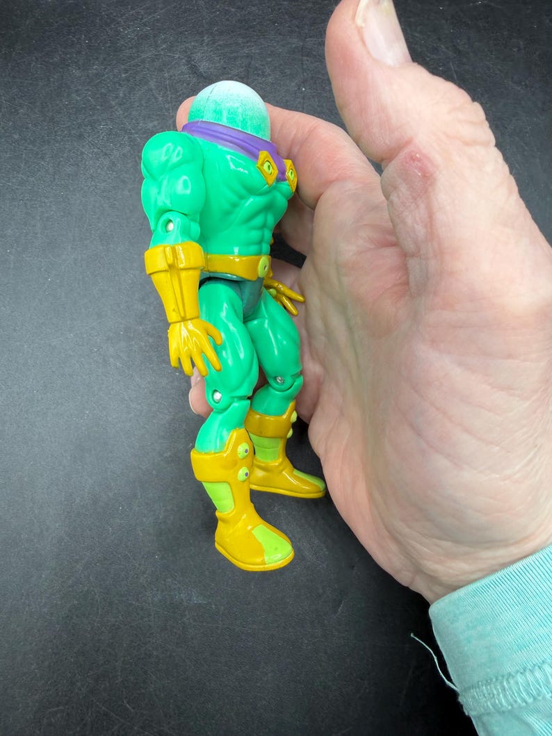 Op de afbeelding: Een groen actiefiguur met een bolvormig hoofd, paarse hoofdband en gele accenten op handschoenen, laarzen en riem. Het figuur wordt in een hand gehouden, wat de grootte en details laat zien. Het speelgoed heeft een gespierde lichaamsbouw en een vintage esthetiek.
