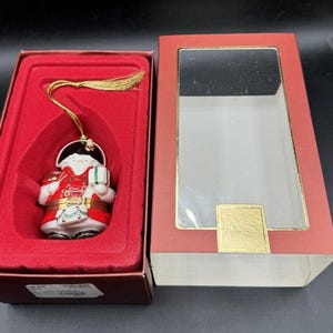 May include: A red and white porcelain Christmas ornament depicting Santa Claus holding a gift box. The ornament is in a red box with a clear plastic window and the word "Lenox" printed on the box.