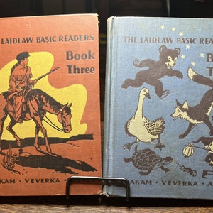 Peut inclure: Deux livres pour enfants vintage, "The Laidlaw Basic Readers" avec des illustrations. Le livre de gauche est intitulé "Book Three" et représente un cow-boy à cheval. Le livre de droite est intitulé "Book Two" et représente un ours, une oie, un renard et une tortue.