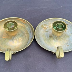Puede incluir: Dos candelabros de latón con base redonda y una pequeña copa redonda para velas. Los candelabros tienen un aspecto desgastado y envejecido.