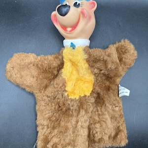 May include: A vintage Yogi Bear hand puppet. The puppet features a tan face with a blue hat, a black nose, and a smiling expression. The body is brown with a yellow scarf and white collar. A tag is attached to the puppet.