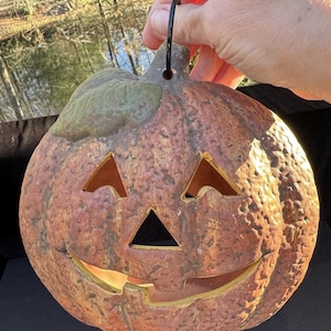 May include: A rustic, orange ceramic jack-o'-lantern with a carved face and a black metal handle. The pumpkin has a textured surface and a green stem. The eyes and mouth are cut out, allowing light to shine through.