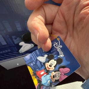 May include: A Disney-branded tag featuring Mickey and Minnie Mouse. The tag has a blue background with a cityscape design. Mickey Mouse is wearing red shorts and yellow shoes, while Minnie Mouse has a pink bow.