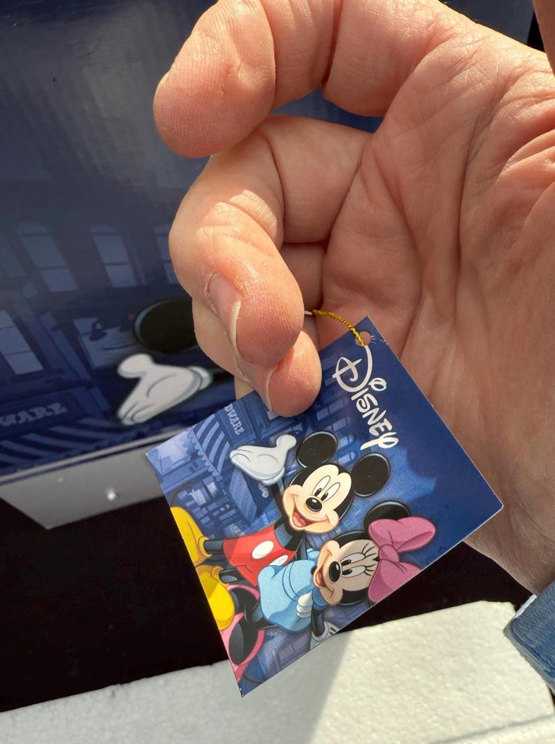 May include: A Disney-branded tag featuring Mickey and Minnie Mouse. The tag has a blue background with a cityscape design. Mickey Mouse is wearing red shorts and yellow shoes, while Minnie Mouse has a pink bow.