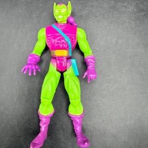 May include: A Green Goblin action figure with a green face and body, wearing a pink top, purple gloves, and boots. The figure has a teal strap across the chest and a brown belt. The figure is a collectible toy.