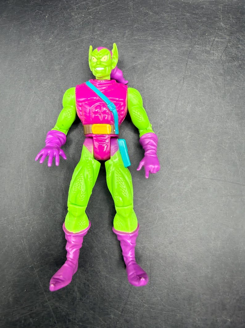 May include: A Green Goblin action figure with a green face and body, wearing a pink top, purple gloves, and boots. The figure has a teal strap across the chest and a brown belt. The figure is a collectible toy.
