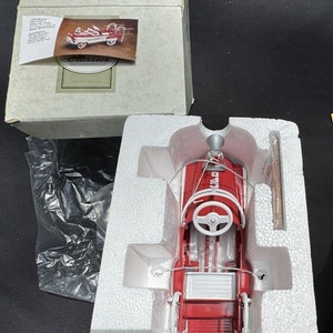 May include: A red and white toy fire truck pedal car with a white steering wheel and a black ladder. The car is in a white foam box with a white plastic bag.