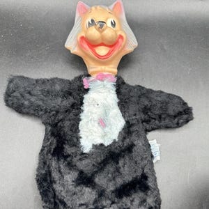 May include: A vintage cat hand puppet with a smiling face and black fur body. The cat's head is tan with pink ears and a red mouth. The puppet has a white and pink collar.