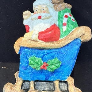 Op de afbeelding: Een kerstornament met de kerstman in een blauwe slee, vergezeld van een teddybeer en een groene zak. De kerstman draagt een rood pak en hoed, met een witte baard. De slee is versierd met hulst en bessen.