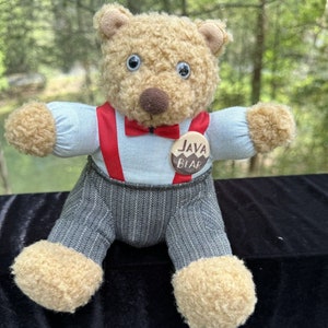 Pode incluir: Um urso de peluche marrom vestindo uma camisa azul com suspensórios vermelhos e uma gravata borboleta vermelha. O urso tem um nariz marrom e grandes olhos pretos. O urso está usando um botão que diz "Java Bear".