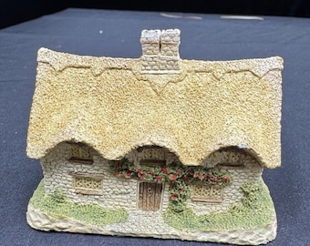 Może przedstawiać: Miniaturowa figurka domku z trzcinowym dachem, kamiennymi ścianami i kominem. Domek posiada drewniane drzwi, okna i pnące czerwone róże. Podstawa pokryta jest zielonymi liśćmi.