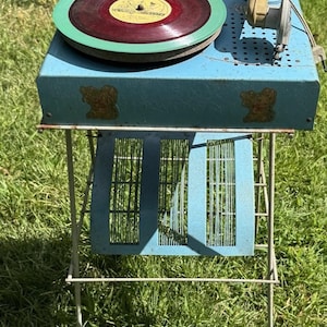 May include: Vintage blue record player with a red vinyl record and green trim. The record player has a tan tone arm and is on a metal stand with a blue cage-like design. The record player is outdoors on a grassy surface.