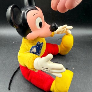 May include: A vintage Mickey Mouse plush doll. The doll is wearing a yellow shirt, red pants, and yellow shoes. Mickey has black ears, a black nose, and white gloves. The doll is sitting down with its arms outstretched.