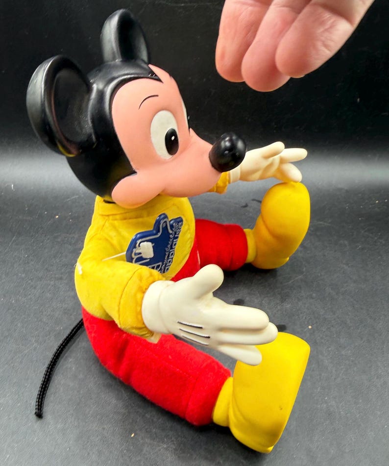 May include: A vintage Mickey Mouse plush doll. The doll is wearing a yellow shirt, red pants, and yellow shoes. Mickey has black ears, a black nose, and white gloves. The doll is sitting down with its arms outstretched.