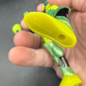 May include: A green and yellow action figure with black accents. The figure's foot is yellow, and the sole is marked with "&copy; 1993 MARVEL MADE IN CHINA". The figure is held in a hand.