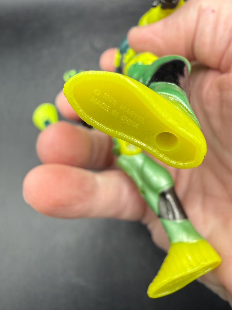 May include: A green and yellow action figure with black accents. The figure's foot is yellow, and the sole is marked with "&copy; 1993 MARVEL MADE IN CHINA". The figure is held in a hand.