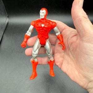 Puede incluir: Figura de acci&oacute;n roja y plateada de un superh&eacute;roe con casco, peto y botas. La figura tiene articulaciones y se sostiene en una mano sobre un fondo oscuro. El dise&ntilde;o de la figura recuerda a un personaje de c&oacute;mic.