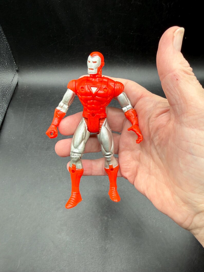 Puede incluir: Figura de acci&oacute;n roja y plateada de un superh&eacute;roe con casco, peto y botas. La figura tiene articulaciones y se sostiene en una mano sobre un fondo oscuro. El dise&ntilde;o de la figura recuerda a un personaje de c&oacute;mic.