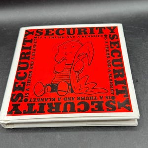 May include: A square, white-edged book with a red center featuring a cartoon illustration of a child clutching a blanket. The text "SECURITY IS A THUMB AND A BLANKET" is printed around the edges of the red square in black.