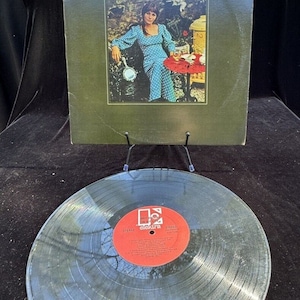 Puede incluir: Una portada de álbum de vinilo y un disco. La portada es verde y presenta a una mujer con un vestido de lunares azules y blancos sentada en una silla. El texto de la portada dice "In My Life / Judy Collins". El disco es negro con una etiqueta roja que dice "In My Life / Judy Collins / Elektra".