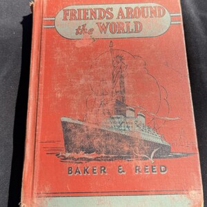 以下が含まれることがあります： 「Friends Around the World」と題されたヴィンテージ本。赤い表紙には、船と自由の女神のイラストが描かれています。タイトルは水色の楕円形で、出版社はBaker & Reedです。