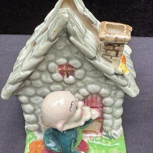 Könnte beinhalten: Keramikfigur eines kleinen Hauses mit Schornstein, roter Tür und steinartiger Textur. Eine kleine Figur in einer grünen Jacke und roter Hose kniet vor der Tür. Das Haus hat ein hellgrünes Dach und Wände.