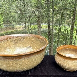 May include: Two ceramic bowls, one large and one small, with a speckled brown and beige glaze. The bowls are sitting on a black surface.