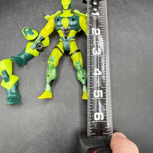May include: A yellow and green action figure with a tape measure next to it. The figure is approximately 4 inches tall. The figure has a helmet, armor, and detached arm. The figure is on a dark surface.