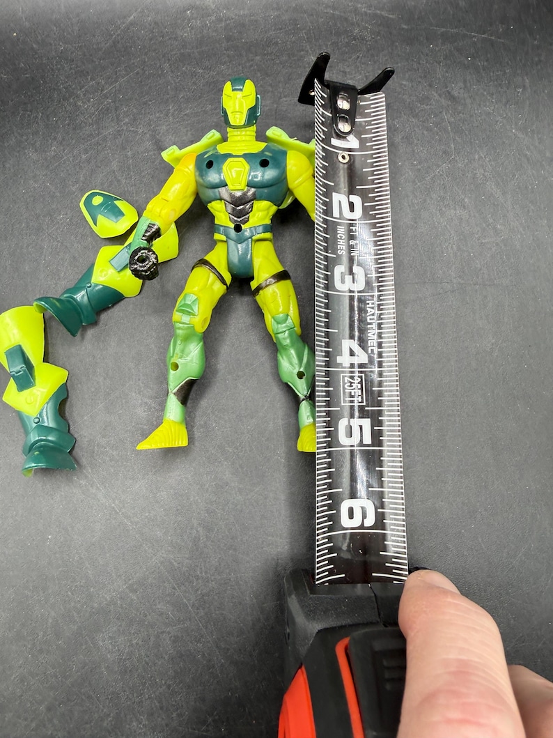 May include: A yellow and green action figure with a tape measure next to it. The figure is approximately 4 inches tall. The figure has a helmet, armor, and detached arm. The figure is on a dark surface.