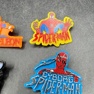 Może przedstawiać: Dwa zawieszki z motywem Spider-Mana. Jeden przedstawia Spider-Mana w czerwono-niebieskim kostiumie z napisem "SPIDER-MAN" w kolorze ż&oacute;łtym. Drugi to zawieszka Cyborg Spider-Man z napisem "CYBORG SPIDER-MAN" w kolorze srebrnym.