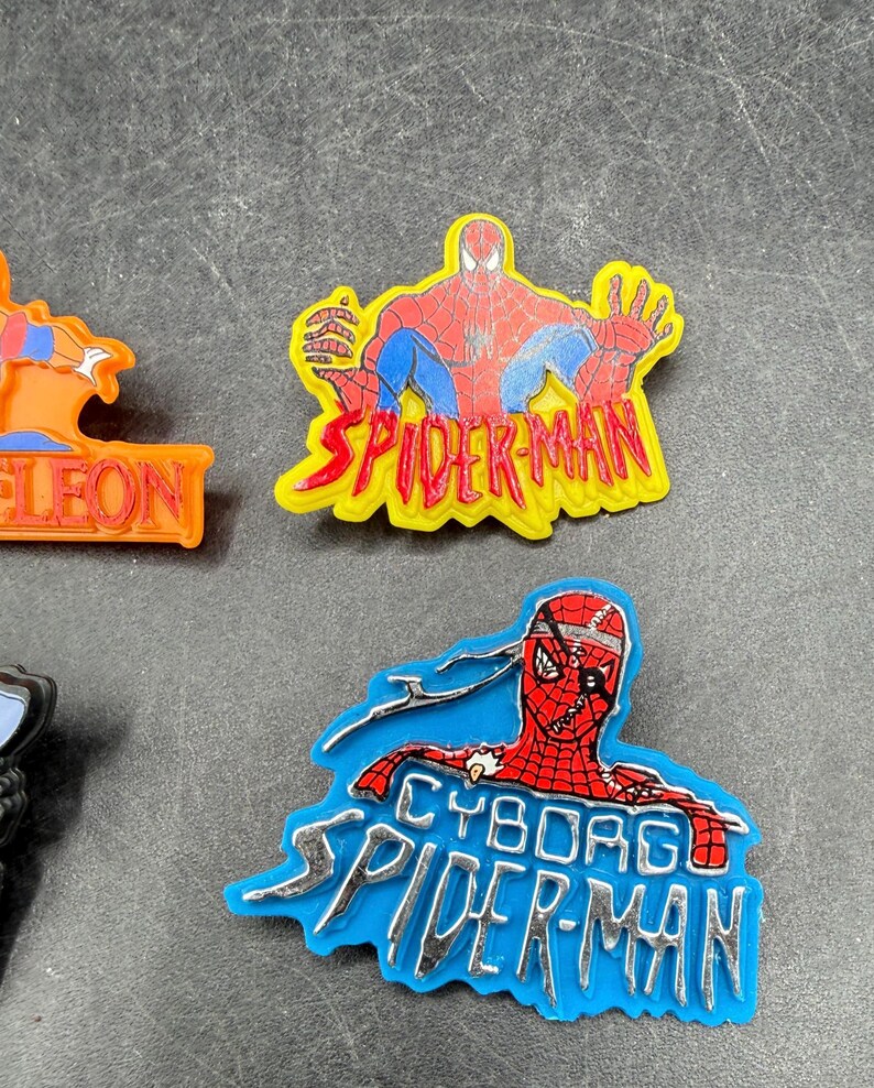 Może przedstawiać: Dwa zawieszki z motywem Spider-Mana. Jeden przedstawia Spider-Mana w czerwono-niebieskim kostiumie z napisem "SPIDER-MAN" w kolorze ż&oacute;łtym. Drugi to zawieszka Cyborg Spider-Man z napisem "CYBORG SPIDER-MAN" w kolorze srebrnym.