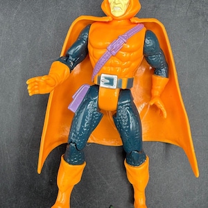 May include: Orange action figure of a character with a hooded cowl and cape. The figure has orange gloves, boots, and a belt with a silver buckle. The figure has a purple belt and a small pumpkin accessory.