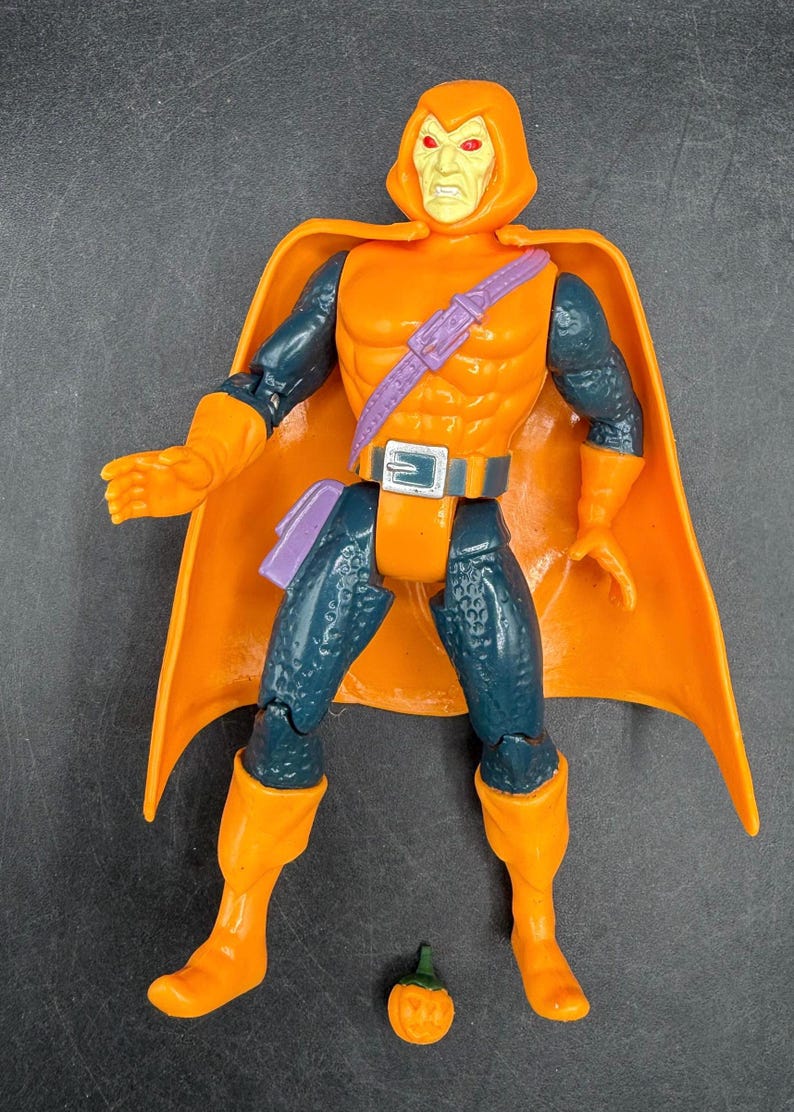 May include: Orange action figure of a character with a hooded cowl and cape. The figure has orange gloves, boots, and a belt with a silver buckle. The figure has a purple belt and a small pumpkin accessory.