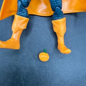 May include: A close-up of a superhero action figure with orange boots and a matching cape. The figure has blue legs and a small, carved orange pumpkin with a green stem is on the surface.