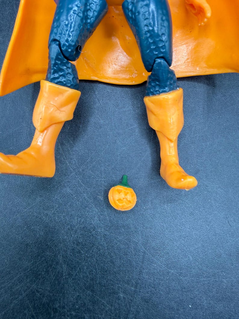 May include: A close-up of a superhero action figure with orange boots and a matching cape. The figure has blue legs and a small, carved orange pumpkin with a green stem is on the surface.