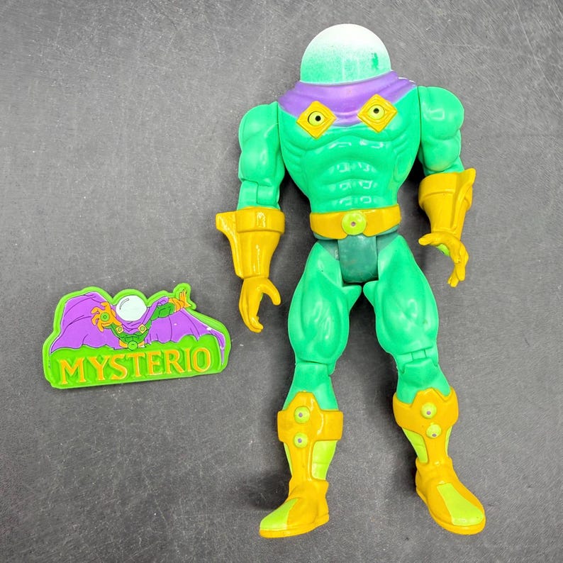 Op de afbeelding: Een groene actiefiguur met gele accenten, een doorzichtige, koepelvormige kop en een paars masker. Daarnaast een groen en paars embleem met het woord "MYSTERIO".