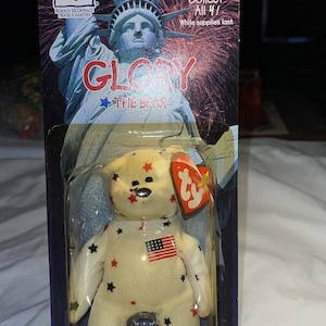 Puede incluir: Un oso de peluche blanco Ty Beanie Baby llamado "Glory" con estrellas rojas y negras y una bandera estadounidense en el pecho. El oso está empaquetado en una caja de plástico con un fondo azul y blanco que presenta la Estatua de la Libertad y fuegos artificiales. El empaque también incluye el logotipo de Ty y el texto "Collect All 4! While supplies last."