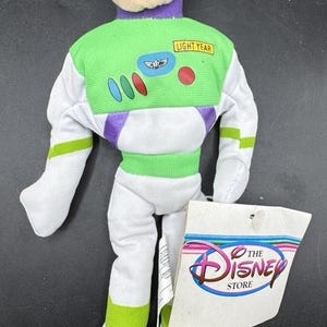 May include: A Buzz Lightyear plush doll from the Disney Store. The doll features a green and white spacesuit with purple accents, and the words "LIGHT YEAR" on the chest. A Disney Store tag is attached.