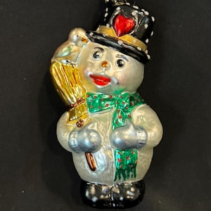 May include: A snowman Christmas ornament with a black top hat featuring a red heart, a green scarf, and a gold star on top. The snowman holds a broom and a bird. The ornament has a silver body and black boots.