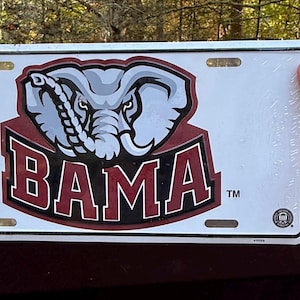 May include: White license plate featuring the Alabama Crimson Tide logo. The logo includes an elephant head in gray and white above the word "BAMA" in a maroon and white font. The plate has four mounting holes.