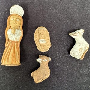 Puede incluir: Conjunto de cuatro pequeñas figuras de cerámica que representan a una mujer, un bebé en un pesebre y dos corderos. Las figuras están pintadas en marrón y blanco.