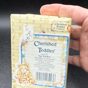 May include: A small, rectangular box with a yellow floral pattern and the words "Cherished Teddies" in blue. The box features an illustration of a teddy bear with a crutch and the text "God Bless Us Everyone".