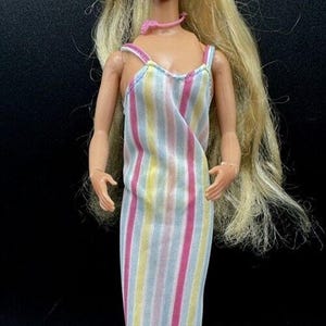 Może przedstawiać: Lalka Barbie vintage z długimi blond włosami, niebieskimi oczami i różowym naszyjnikiem. Ma na sobie sukienkę w paski w kolorze różowym, żółtym, niebieskim i białym.