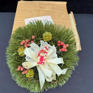 May include: A small, round Christmas wreath made of green faux pine needles, decorated with red berries, small pinecones, and a cream-colored bow. A small figurine of a child holding a candy cane is attached to the bow.