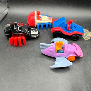 May include: Four toy vehicles featuring Spider-Man characters. The toys include a black car with red accents, a red and blue car, a red and blue vehicle with a trailer, and a pink and blue aircraft. Each vehicle has a Spider-Man figure.